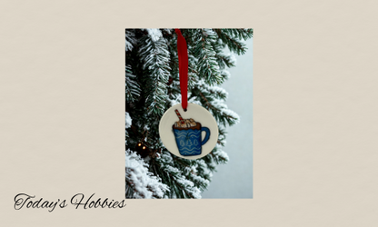 Handcrafted Christmas ornament for home featuring a sublimation glass design of a blue mug with whipped cream hanging on a snowy pine tree